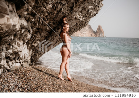 A tourist woman in bikini on beach enjoys the turquoise sea during her summer holiday. Beach vacation. Woman with braids dreadlocks standing with her arms raised near basalt rock enjoying beach ocean A tourist woman in bikini on beach enjoys the turquoise sea during her summer holiday. Beach vacation. Woman with braids dreadlocks standing with her arms raised near basalt rock enjoying beach ocean 111984910