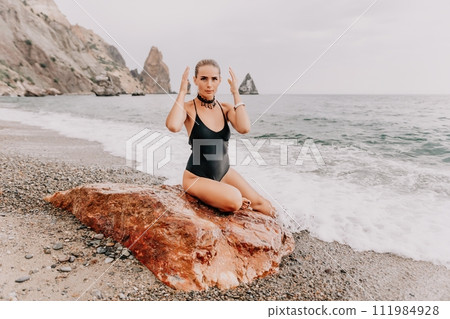 Woman travel portrait. Happy woman with long hair looking at camera and smiling. Close up portrait cute woman in black bikini posing on a red volcanic rock on the sea beach 111984928