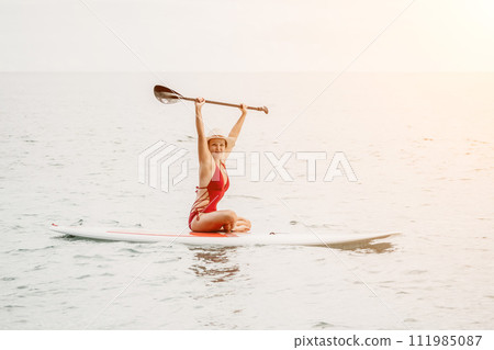 Woman in red bikini on sup board. Happy lady with blond hair in red bathing suit chilling and sunbathing by turquoise sea ocean on sunset. Holiday, vacation and recreational concept. Woman in red bikini on sup board. Happy lady with blond hair in red bathing suit chilling and sunbathing by turquoise sea ocean on sunset. Holiday, vacation and recreational concept. 111985087