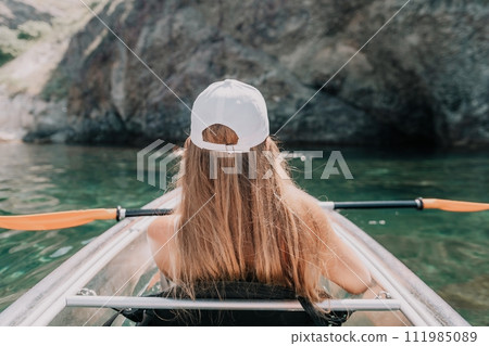 Woman in kayak back view. Happy young woman with long hair floating in transparent kayak on the crystal clear sea. Summer holiday vacation and cheerful female people relaxing having fun on the boat Woman in kayak back view. Happy young woman with long hair floating in transparent kayak on the crystal clear sea. Summer holiday vacation and cheerful female people relaxing having fun on the boat 111985089
