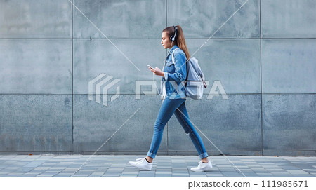 Full length side student woman walking with smartphone and earphones. Generative AI 111985671