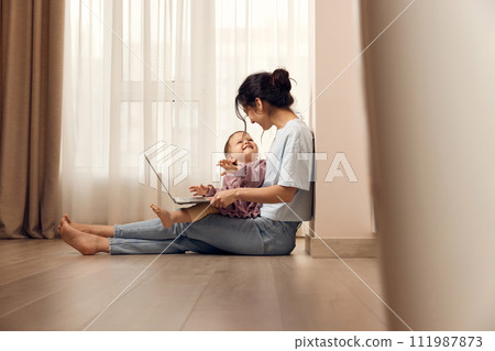 beautiful casual woman sitting on the floor and working on laptop with her little child girl at home 111987873