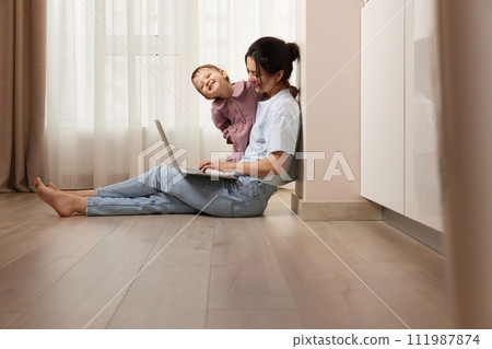 happy casual woman sitting on the floor and working on laptop with her little baby girl at home happy casual woman sitting on the floor and working on laptop with her little baby girl at home 111987874