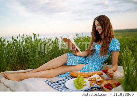Young woman sits on a plaid with a book. Summer picnic in nature. Healthy food. 111988800