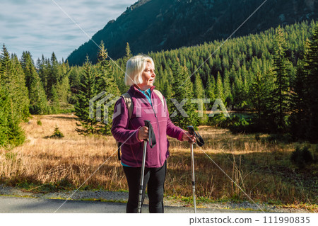 Mature caucasian blonde woman on vacation, having a hike in autumn mountains with backpack and hiking poles, enjoying adventure. 111990835