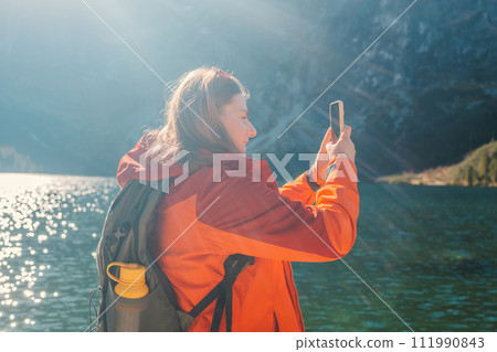 Rear shot of traveler woman in sportswear standing on the rocky shore and making selfie on smart phone or video. Taking photo of green hills and mountains on Morskie Oko lake. Hiking travel and Rear shot of traveler woman in sportswear standing on the rocky shore and making selfie on smart phone or video. Taking photo of green hills and mountains on Morskie Oko lake. Hiking travel and 111990843