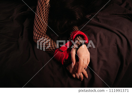 female hands in red leather BDSM handcuffs on a black sheet in close up. Submissive woman slave girl is lying on bed in bedroom. Sex with submission and domination female hands in red leather BDSM handcuffs on a black sheet in close up. Submissive woman slave girl is lying on bed in bedroom. Sex with submission and domination 111991371