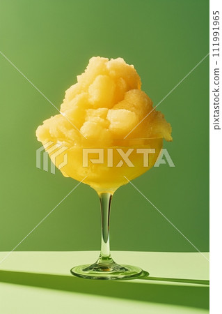 A vivid yellow slushie in a clear stemmed glass, on a green background with a strong shadow 111991965