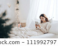 AI-generated content. A young woman in white clothes sits in bed and looks at a mobile phone or tablet. Christmas atmosphere 111992797
