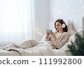 AI-generated content. A young woman in white clothes and headphones lies in bed and listens to music, an audiobook or watches a movie. Christmas atmosphere 111992800