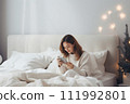 AI-generated content. A young woman in white clothes sits in bed and looks at her mobile phone. Christmas atmosphere 111992801