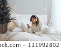 AI-generated content. A young woman in white clothes and headphones lies in bed and listens to music, an audiobook or watches a movie. Christmas atmosphere 111992802