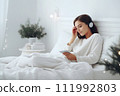 AI-generated content. A young woman in white clothes and headphones lies in bed and listens to music, an audiobook or watches a movie. Christmas atmosphere 111992803