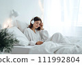AI-generated content. A young woman in white clothes lies in bed and listens to music on headphones. Christmas atmosphere 111992804