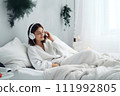 AI-generated content. A young woman in white clothes lies in bed and listens to music on headphones. Christmas atmosphere 111992805