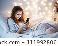 AI-generated content. A teenage girl sits in bed in a bedroom decorated with Christmas decor and looks at a mobile phone or tablet. 111992806