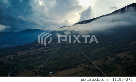 The image shows a towering mountain with jagged peaks, shrouded in wispy clouds against a blue sky. The clouds create a dramatic contrast against the rocky terrain, enhancing the rugged beauty of the 111994375