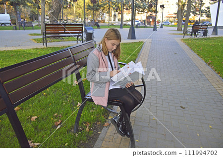 girl studying a book in English on a bench in a city park. additional education 111994702
