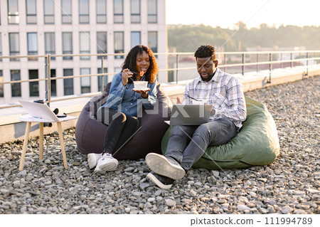 African man and woman working on laptop while sitting at rooftop workplace. 111994789
