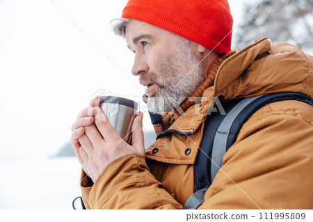Bearded gray-haired man in red hat with thermos in hands in a winter forest 111995809