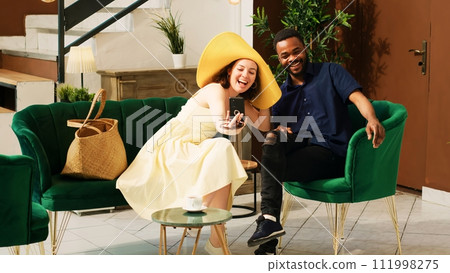 Diverse couple using videocall in hotel lobby, talking to people on online teleconference in lounge area before check in. Tourists enjoying remote video conversation at tropical seaside resort. 111998275
