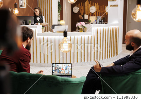 Businessman on videocall in hotel lobby, attending important online conference while they are waiting to check in. Young corporate team talking to executive board on videoconference. Businessman on videocall in hotel lobby, attending important online conference while they are waiting to check in. Young corporate team talking to executive board on videoconference. 111998336