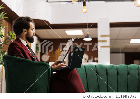Business professional review papers in hotel lobby, waiitng to see room and check in. Entrepreneur travelling for important conference, trying to create new partnership. Front desk reception. 111998355