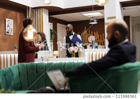 Bellhop filling in registration files for businessman arriving at hotel resort, receiving luxury concierge services. African american employee ensuring relaxing stay for guest. 111998361