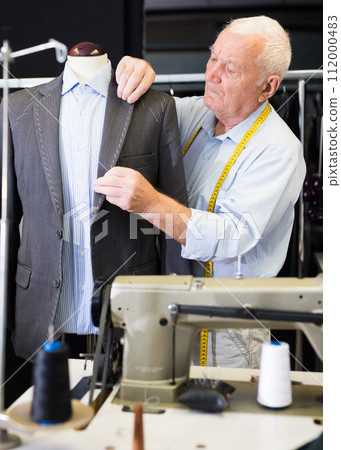 Mature tailor sews a new jacket on a mannequin in sewing studio 112000483