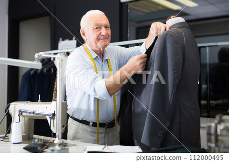 Experienced tailor threading jacket before starting work in workshop Experienced tailor threading jacket before starting work in workshop 112000495