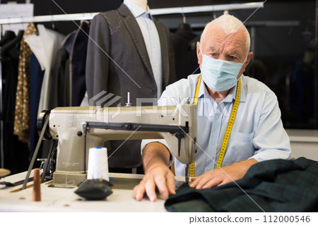 Portrait of elderly tailor in protective mask working on sewing machine 112000546