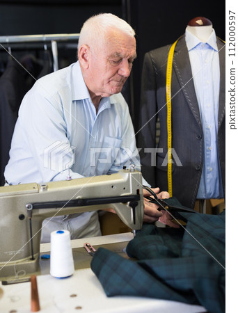Elderly tailor cuts fabric with scissors at workplace in workshop Elderly tailor cuts fabric with scissors at workplace in workshop 112000597