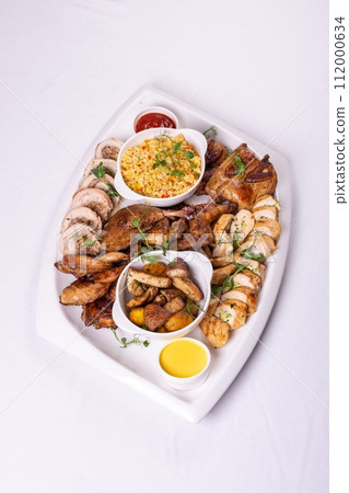 Assorted roasted meats, colorful vegetables, and golden potatoes garnished with fresh parsley and savory sauces on a white platter 112000634