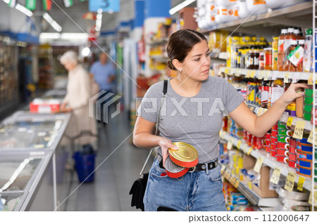 Young woman purchaser choosing tinned fish in big supermarket 112000647