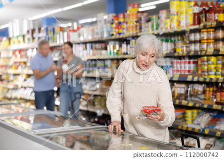 Mature woman choosing frozen food in supermarket. Young woman purchasing goods in grocery store 112000742