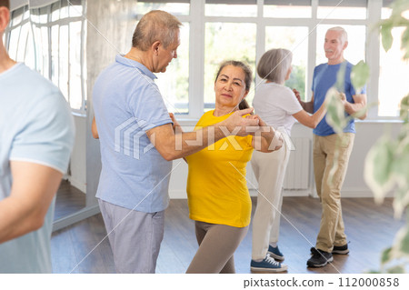 Positive senior woman and man practicing tango dance moves as a couple during a group celebration in dance studio Positive senior woman and man practicing tango dance moves as a couple during a group celebration in dance studio 112000858