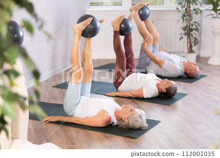 Mature woman doing exercises with pilates ball during group training at gym indoor 112001035