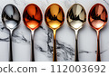 A row of reflective spoons with colored coatings on a marbled white background. 112003692