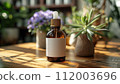 An essential oil bottle with a white label stands on a sunlit wooden table, surrounded by houseplants. 112003696