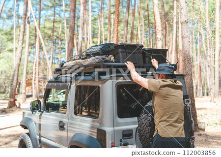 Male camper unloads camping gear from car roof rack at campsite Male camper unloads camping gear from car roof rack at campsite 112003856