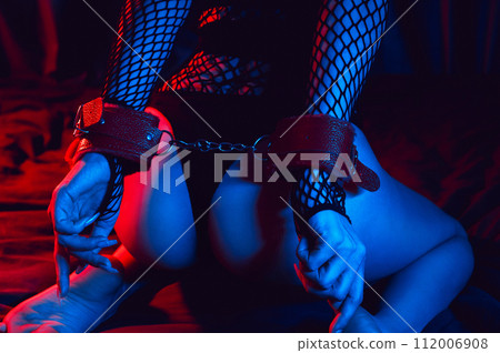 Sexy big ass of a submissive woman slave with handcuffed hands for BDSM sex with submission and domination. Girl in panties is sitting on a bed with neon light 112006908