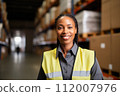 Professional female logistic manager in warehouse. Black woman in high-visibility jacket, overseeing warehouse operations. Strategic planning required to efficiency and meet demands in real-time 112007976