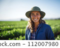 Joyful female farmer in green field. Radiant smiling woman with hat in organic agriculture. Joy from working closely with earth, nurturing growth and being part of the cycle of life. 112007980