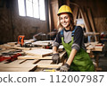 Smiling woman woodworker carpenter in protective gear works at workshop. Craftsmanship and Job Satisfaction, creating with one's hands, love of craft and positive work environment. 112007987