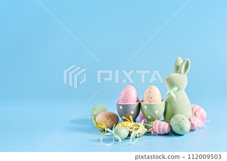 Happy Easter greeting card. soft pastel green Easter bunny figurine and collection of colorful, decorated Easter eggs against a vibrant blue background. Happy Easter greeting card. soft pastel green Easter bunny figurine and collection of colorful, decorated Easter eggs against a vibrant blue background. 112009503