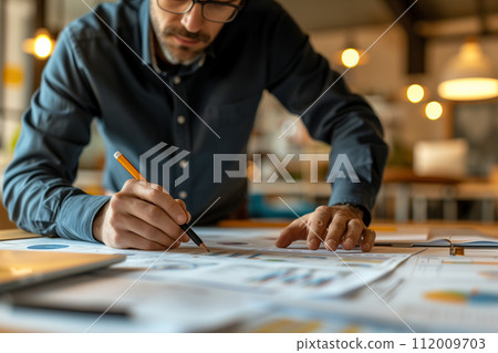 Focused designer with pen in hand closely reviewing detailed project drafts spread across the table. Focused designer with pen in hand closely reviewing detailed project drafts spread across the table. 112009703