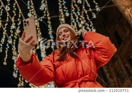 Low-angle view of young cheerful young woman in winter casual clothes making selfie picture, talking video call using holding mobile phone looking to screen, on background festive light illumination. Low-angle view of young cheerful young woman in winter casual clothes making selfie picture, talking video call using holding mobile phone looking to screen, on background festive light illumination. 112010751