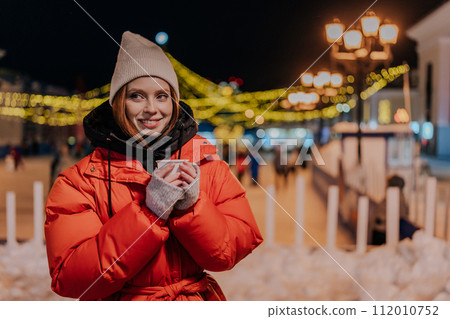 Cheerful young woman wearing warm winter clothes enjoying drinking takeaway cup of coffee outdoors on cold street at night with festive bright illumination. Pretty female walking on urban cold park Cheerful young woman wearing warm winter clothes enjoying drinking takeaway cup of coffee outdoors on cold street at night with festive bright illumination. Pretty female walking on urban cold park 112010752