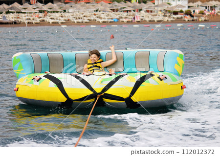 Young boy exudes joy while riding an inflatable tube towed by a boat in the ocean. Happy school child having fun in adventure water park on the sea. 112012372