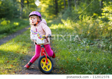 Child riding balance bike. Kids on bicycle in sunny forest. Little girl enjoying to ride glider bike on warm day. Preschooler learning to balance on run bicycle in safe helmet. Sport activity 112012378